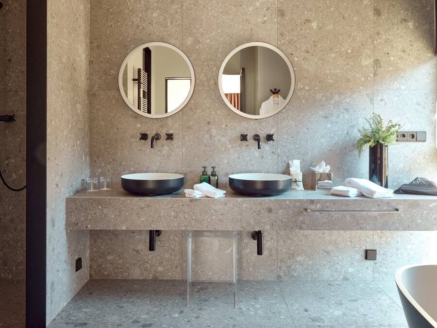 Un bany modern amb dos miralls rodons, un lavabo doble amb una tassa gran, una prestatgeria de vidre transparent, un penjador de tovalloles i una planta en test.