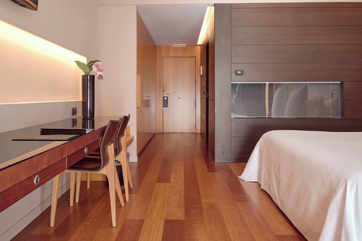 Ein modernes Hotelzimmer mit einem Holztisch, einem Stuhl, einem Bett und einem großen Fenster mit Jalousien.