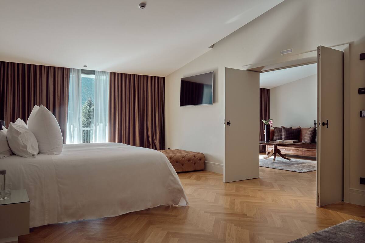 Ein modernes Schlafzimmer mit einem großen Bett, einem an der Wand montierten Fernseher und Blick auf einen Wohnbereich durch eine offene Tür.