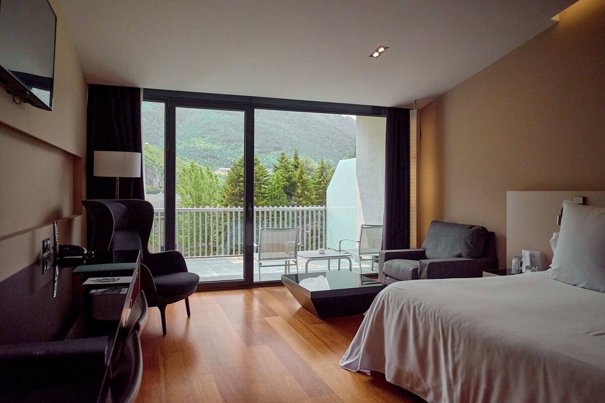 Ein modernes Hotelzimmer mit einem großen Bett, einem Schreibtisch, einem Stuhl und einem Balkon mit Blick auf Grünflächen und Berge.