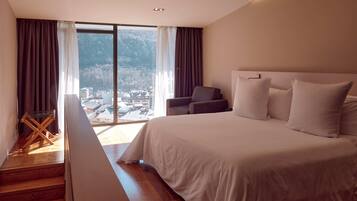 Un dormitorio con una cama grande, una silla y vistas a un paisaje urbano a través de amplios ventanales.
