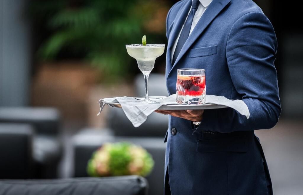 A person in a blue suit holding a tray with a martini glass and a glass of red drink.