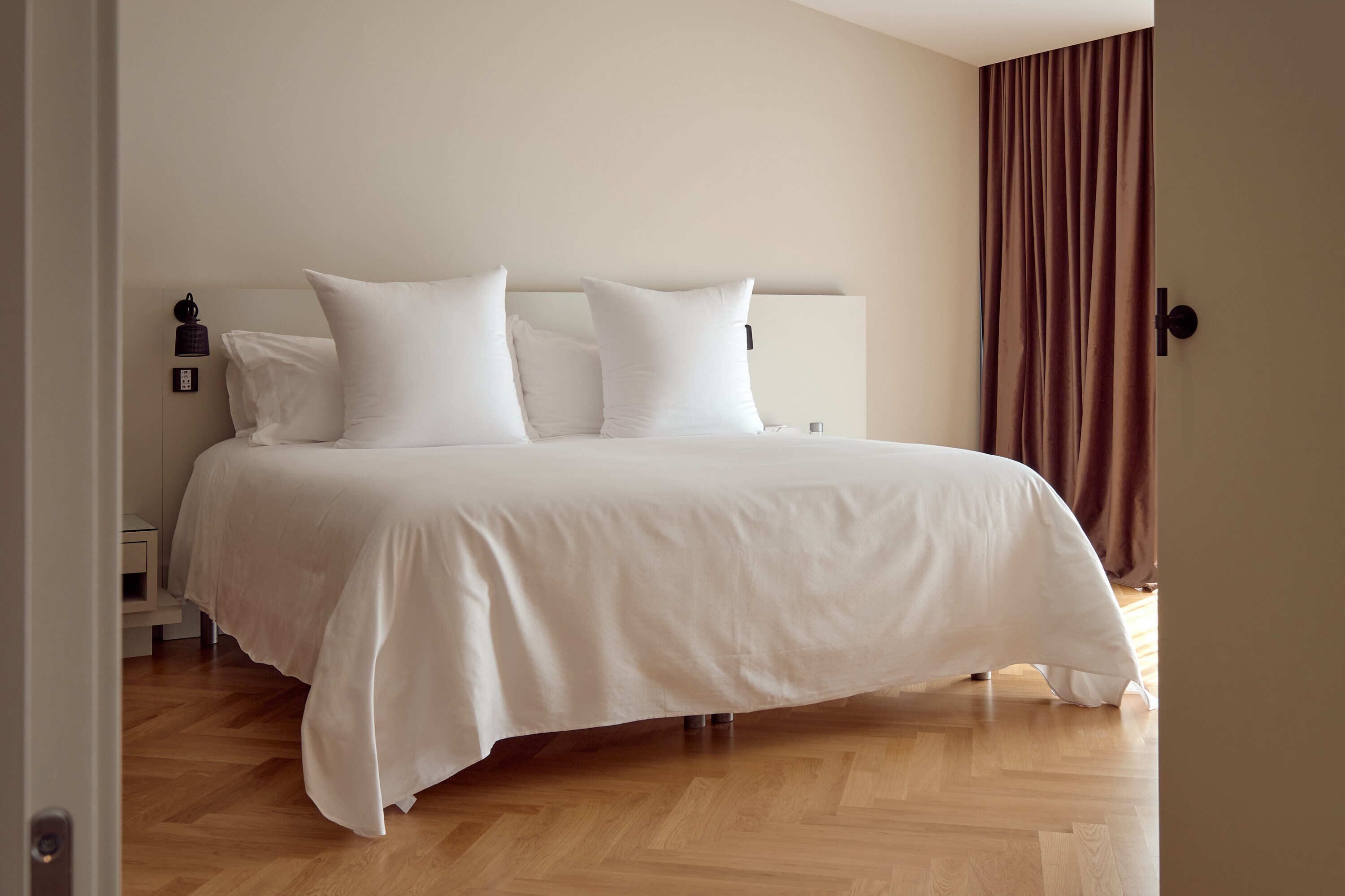 Een netjes opgemaakt bed met witte lakens in een kamer met houten vloeren en een bruin gordijn aan de rechterkant.