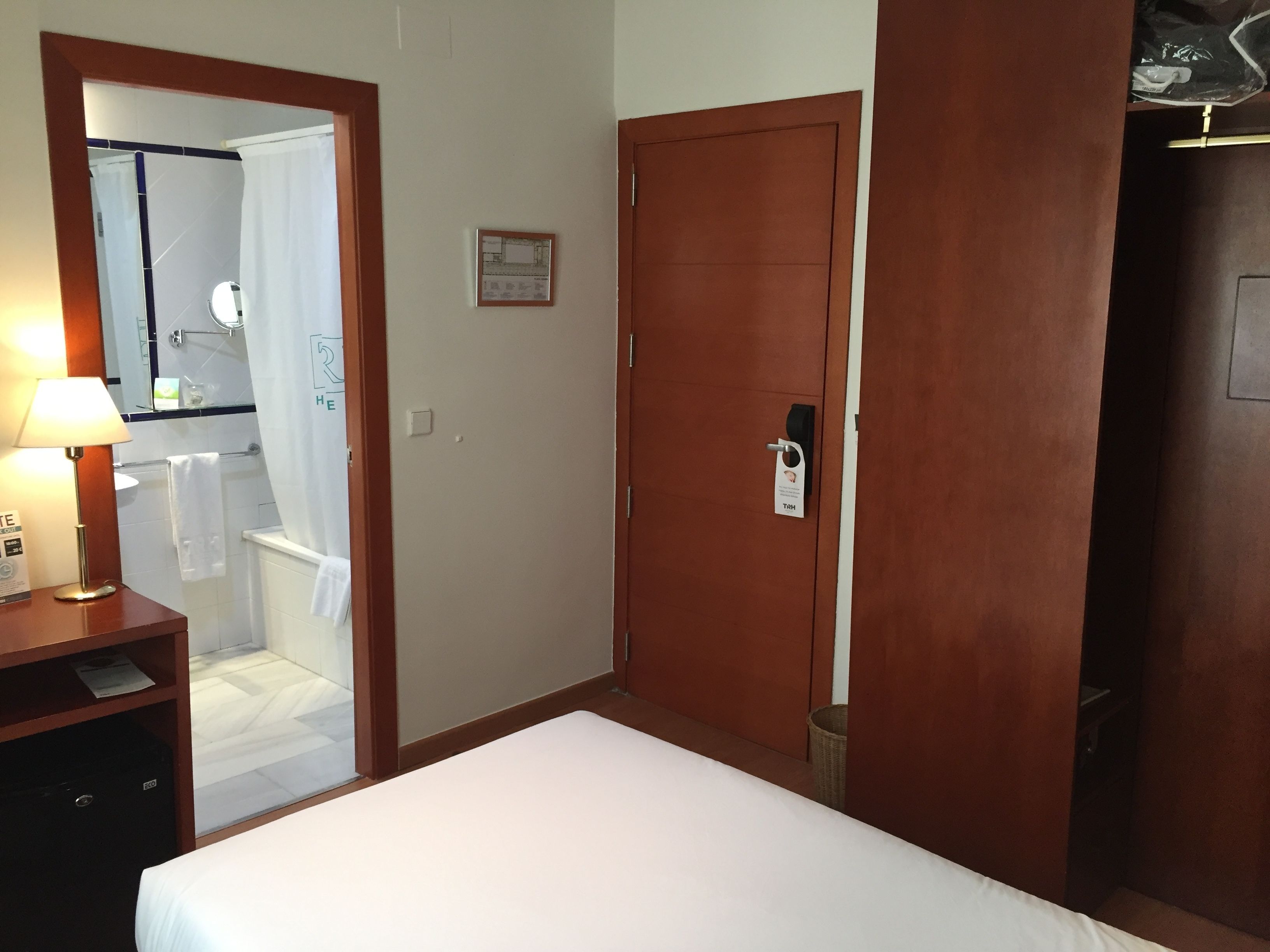 A hotel room with a bed, a wooden wardrobe, a mirror, and a bathroom visible through an open door.