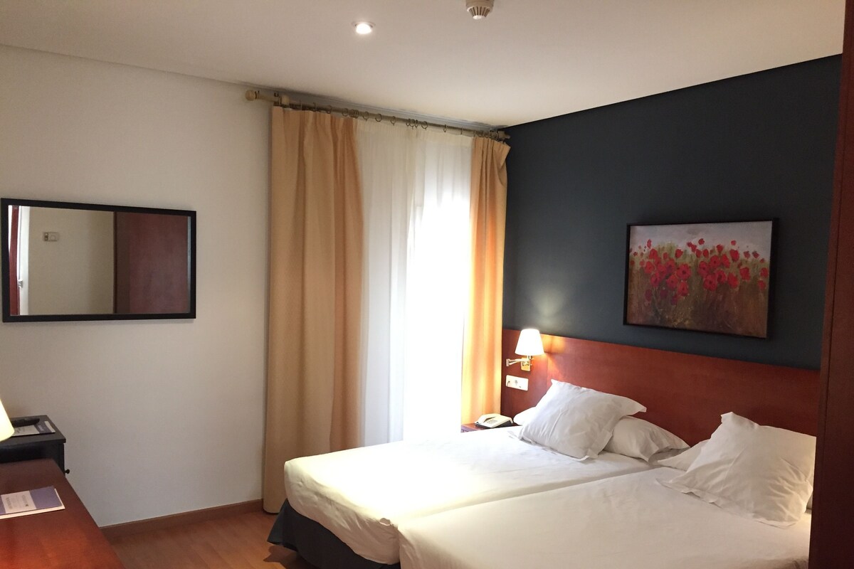 A hotel room with two beds, a desk, a chair, a lamp, a painting, and a mirror.