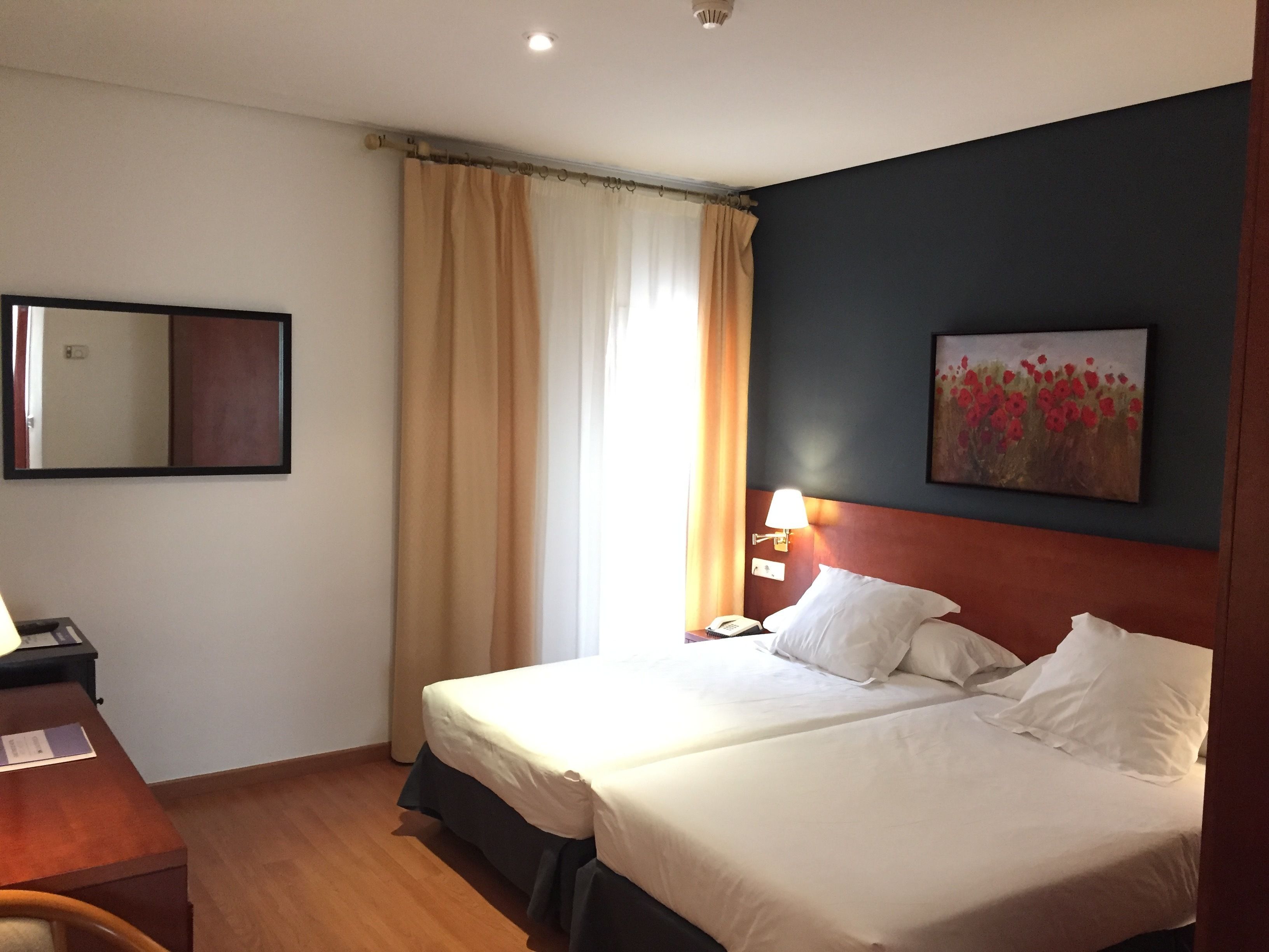 A hotel room with two beds, a desk, a chair, a lamp, a painting, and a window with curtains.