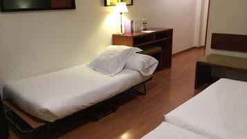 A single bed with a white mattress and pillows, a wooden desk with a lamp, and a small bench.