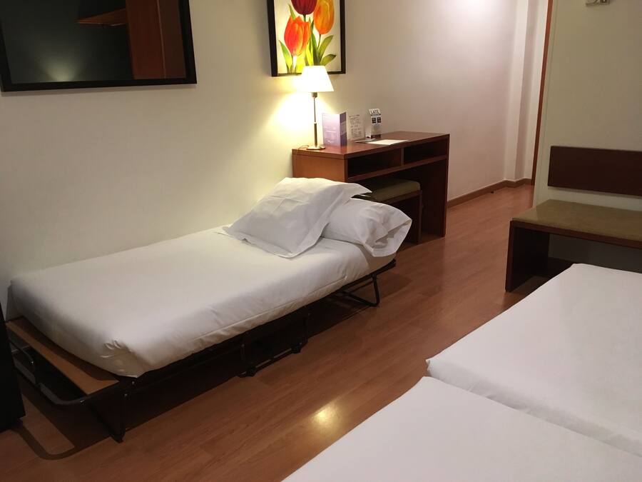 A single bed with a white mattress and pillows, a wooden desk with a lamp, and a small bench.
