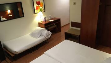 A hotel room with two single beds, a wooden nightstand with a lamp, a small desk, and a chair.