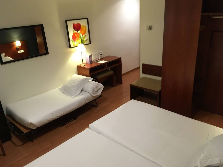 A hotel room with two single beds, a wooden nightstand with a lamp, a small desk, and a chair.