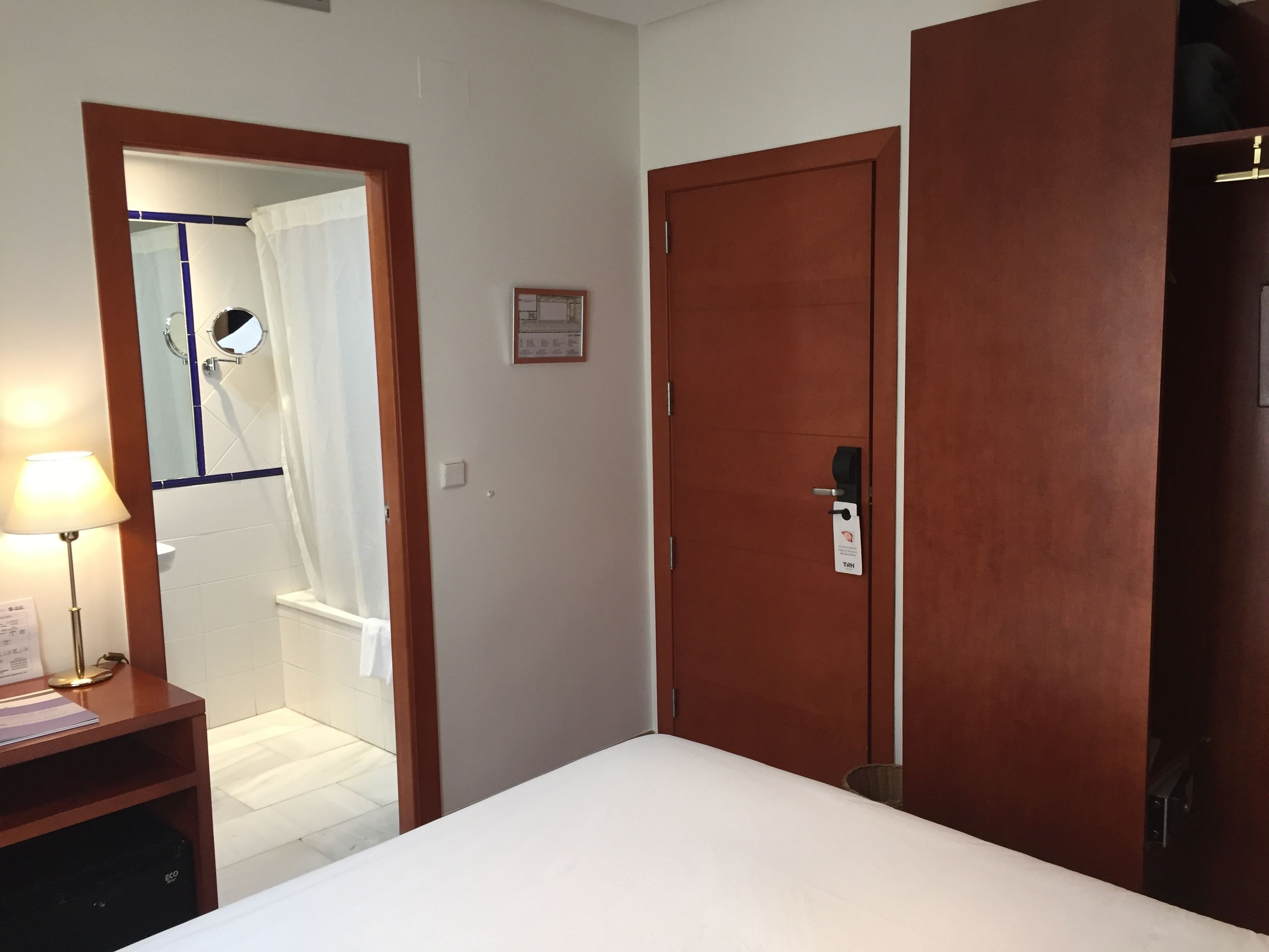 Una camera d'albergo con un letto, una scrivania con una lampada, un angolo doccia e porte in legno.