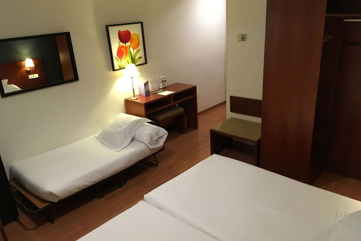 A hotel room with two beds, a desk with a lamp, a chair, a small table, and a wall-mounted painting.