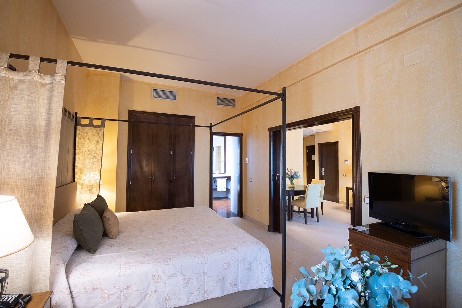Une chambre d’hôtel avec un lit à baldaquin, une télévision et un coin repas visible à travers une porte ouverte.