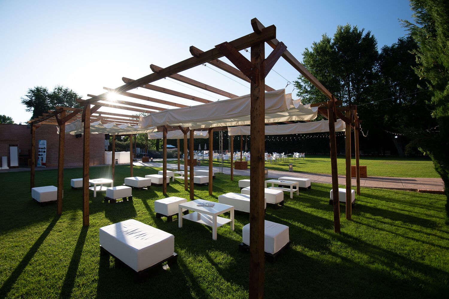 An outdoor patio area with wooden pergolas, white cushioned seating, and a grassy lawn.