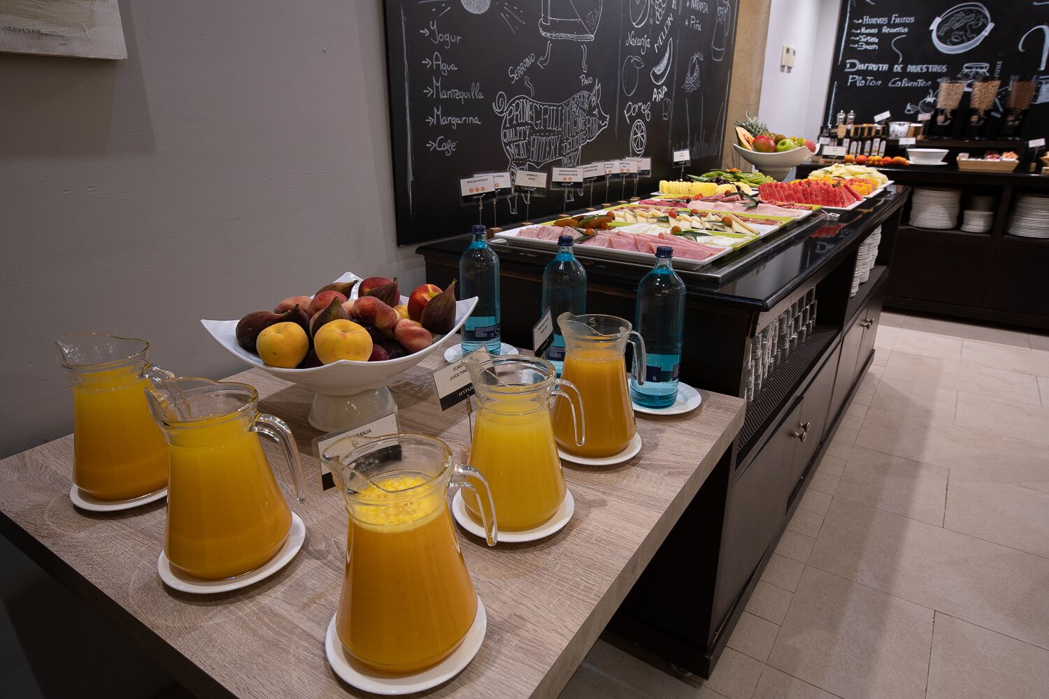 Un bufé de desayuno con diversas frutas, jugos y un menú en una pizarra.
