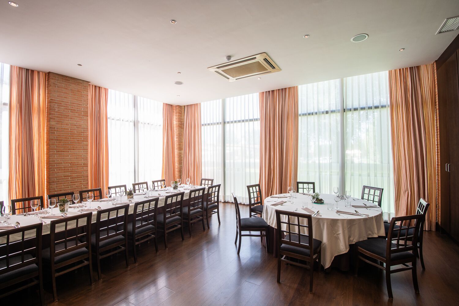 A dining area with multiple tables and chairs, large windows with curtains, and a ceiling vent.