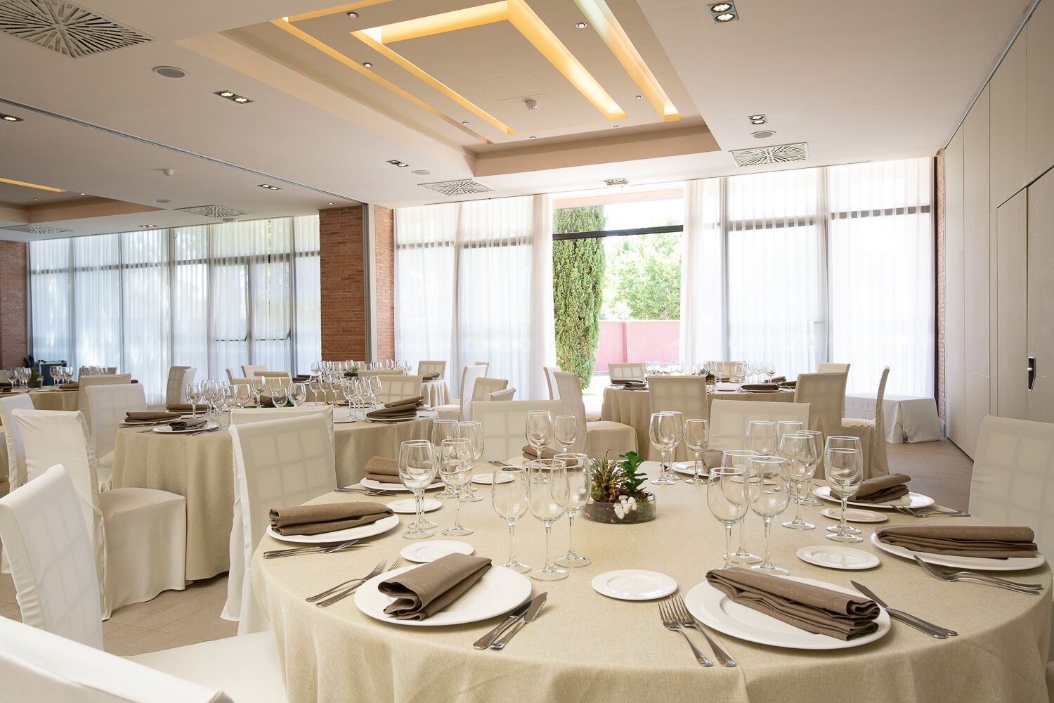 Un comedor amplio con mesas redondas dispuestas para una comida, ventanas grandes que brindan luz natural y vistas al exterior con vegetación.