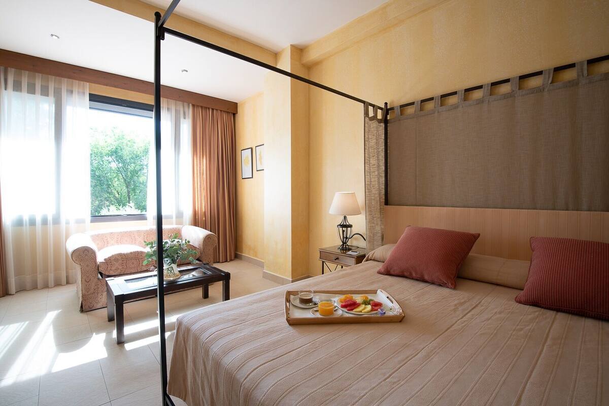A hotel room with a bed, a sitting area with a sofa and a coffee table, a nightstand with a lamp, and a window with curtains.