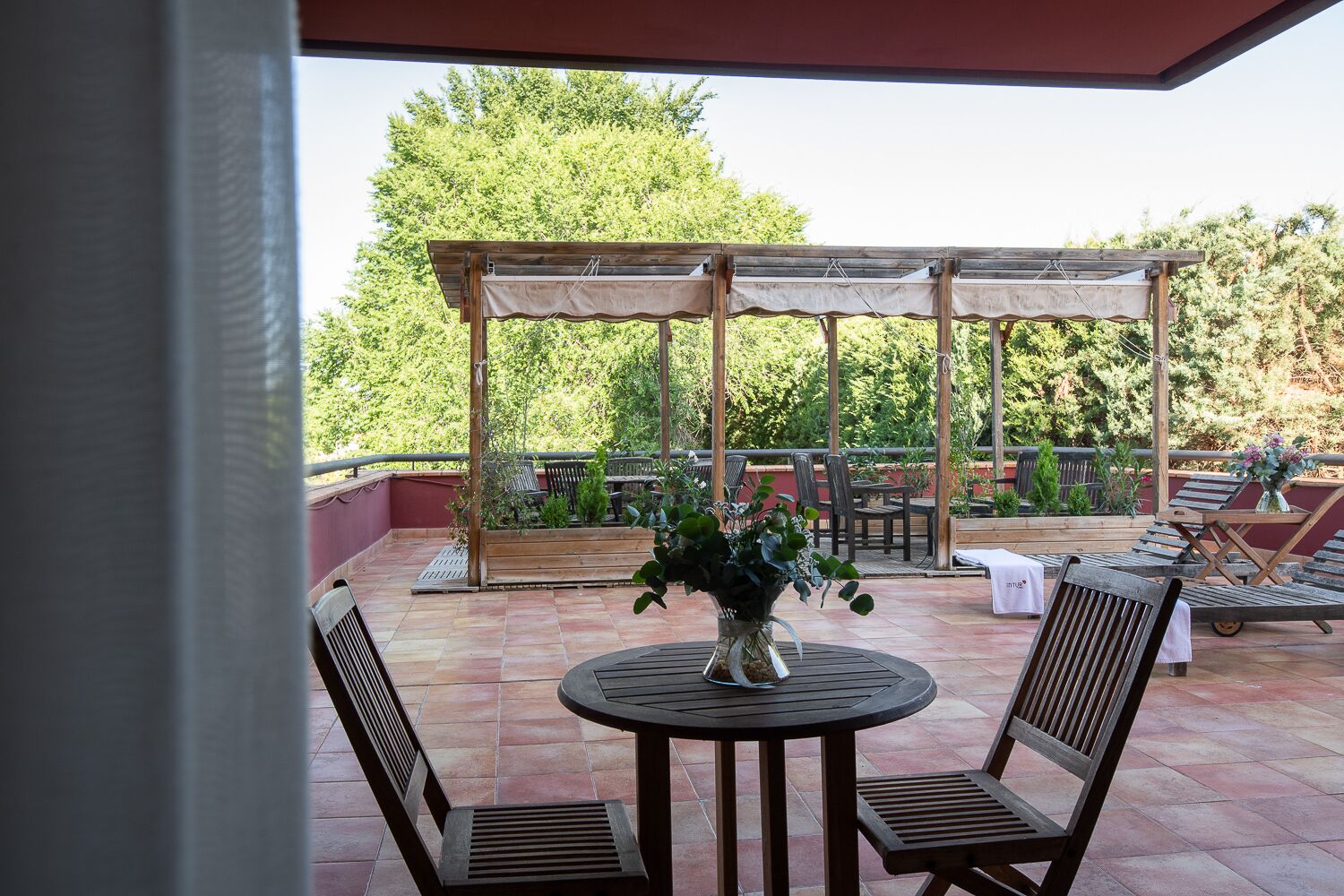 A covered outdoor seating area with a round table and chairs, surrounded by a pergola and a view of trees.