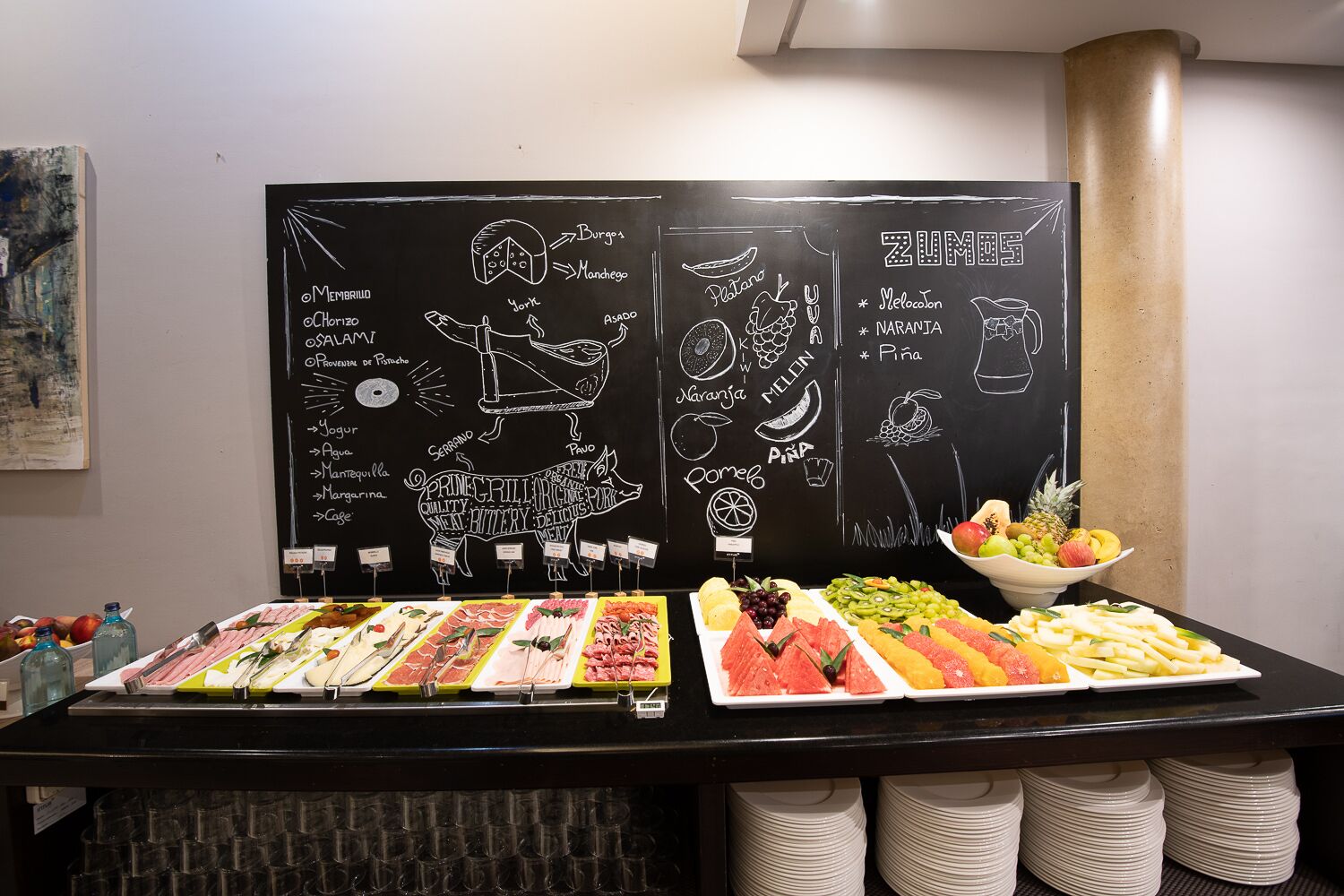 Een buffet met diverse gerechten en een krijtbord waarop gerechten staan vermeld zoals 'Pasta', 'Varkensvlees' en 'Pasta Primavera'.