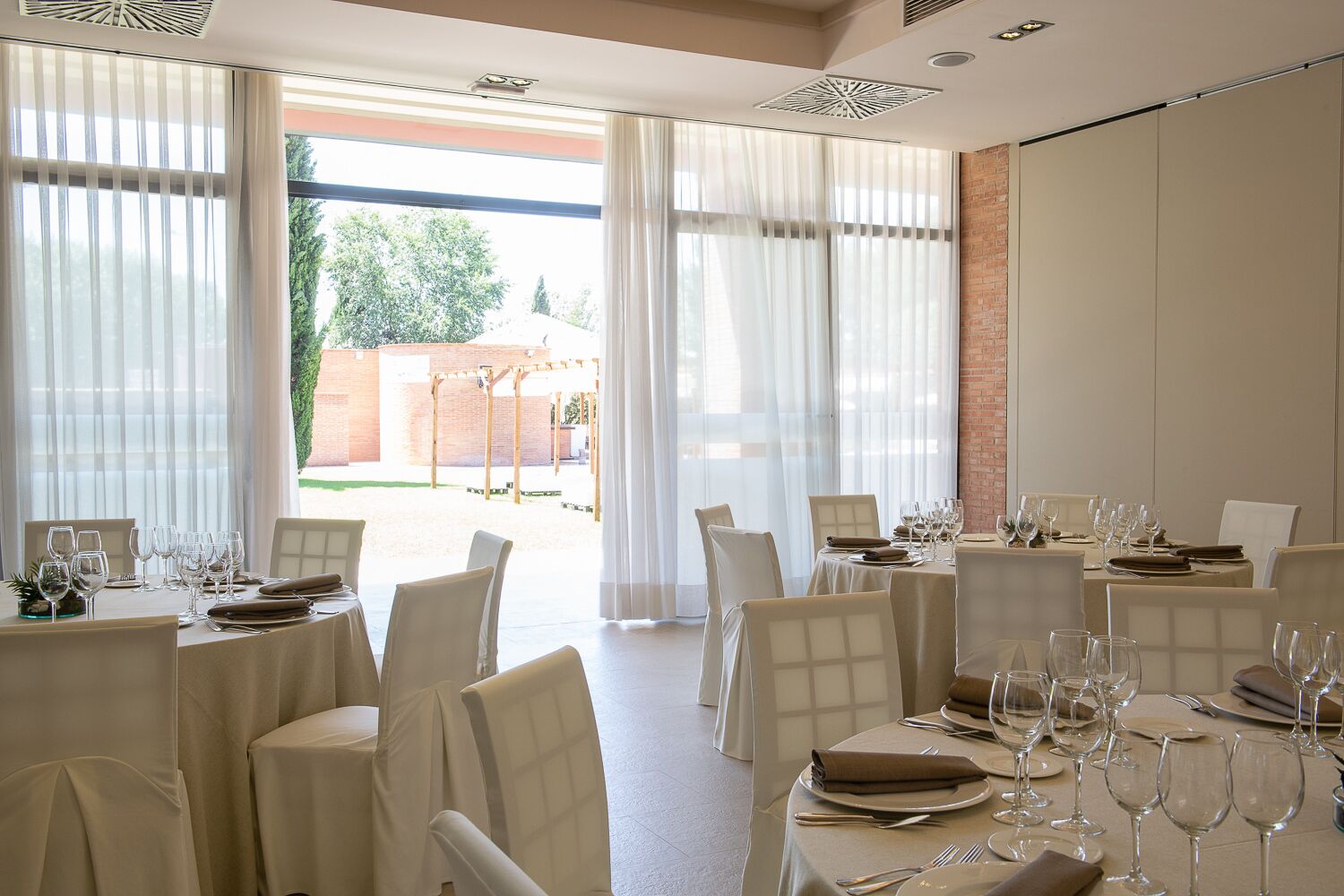 A dining area with tables set for a meal, large windows with sheer curtains, and a view of a brick wall and greenery outside.