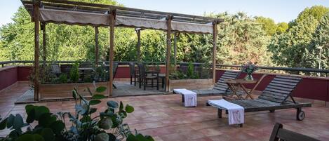 An outdoor patio with wooden furniture, a pergola, and a view of trees.