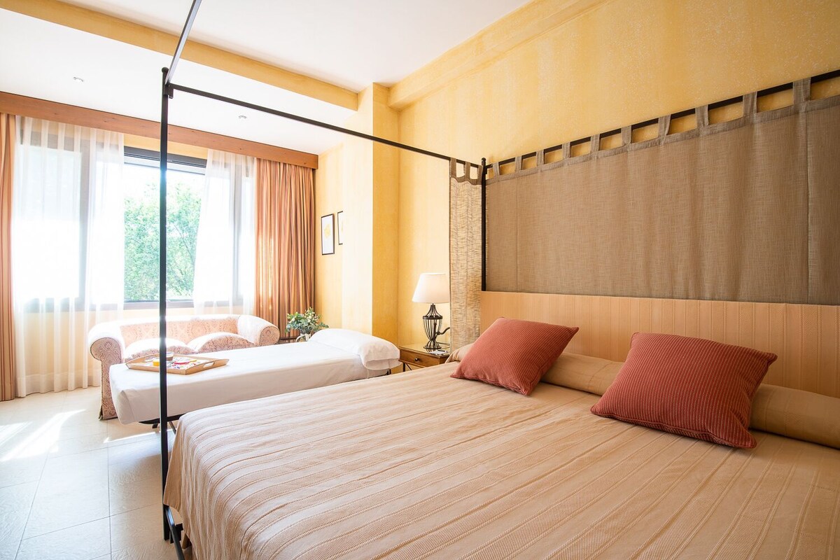 A hotel room with a large bed, a smaller bed, a canopy bed, a nightstand, a lamp, and a window with curtains.
