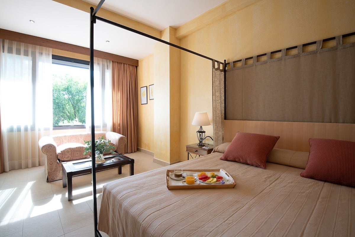 A hotel room with a bed, a sitting area with a sofa and a coffee table, a nightstand with a lamp, and a window with curtains.