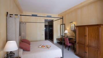 Habitación de hotel con cama con dosel, un escritorio con silla, armario y baño visible a través de una puerta abierta.