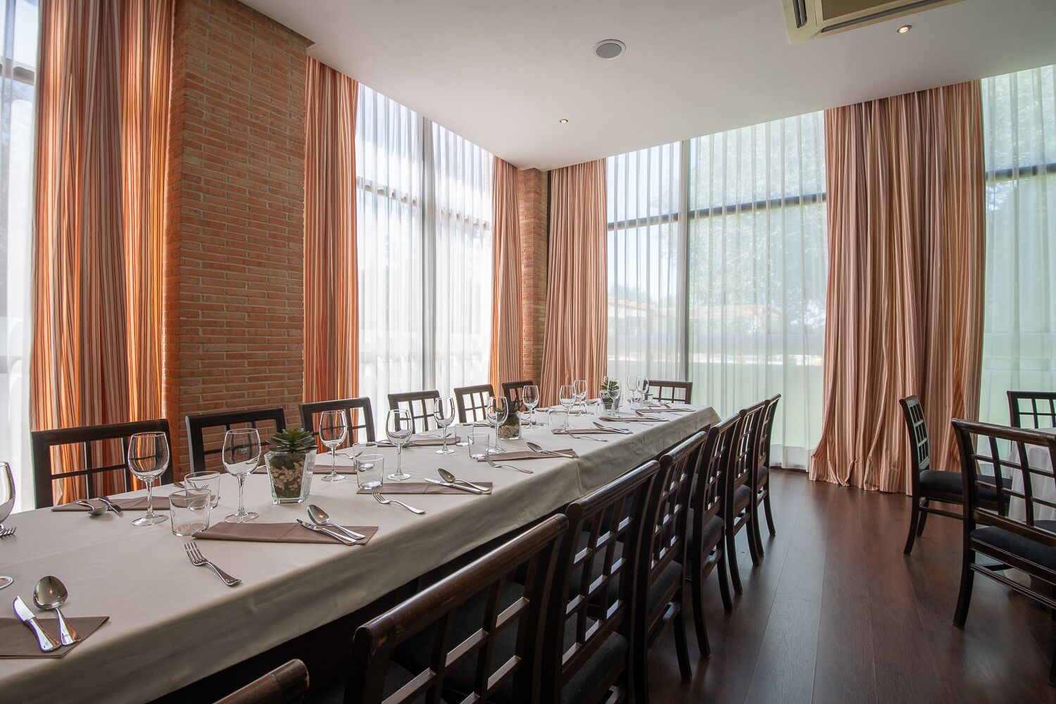 Una larga mesa de comedor preparada para una comida en una habitación con amplios ventanales y paredes de ladrillo.