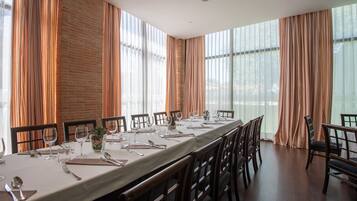 Una larga mesa de comedor preparada para una comida en una habitación con amplios ventanales y paredes de ladrillo.