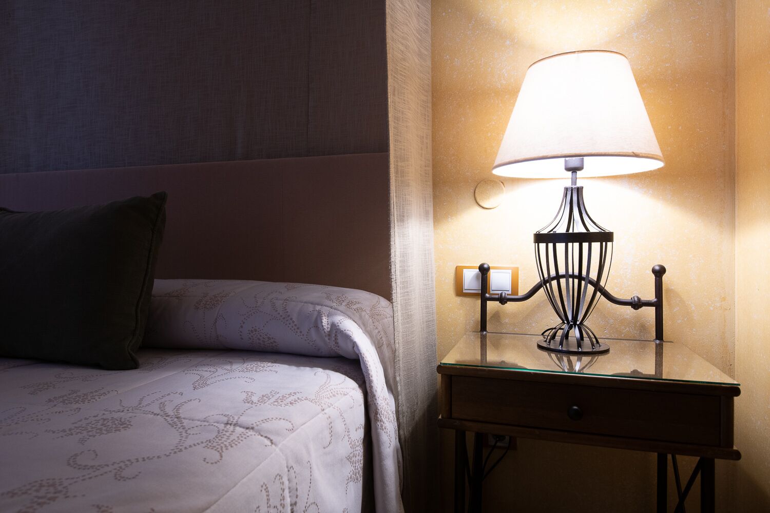 A neatly made bed with a patterned bedspread and a single green pillow. A table lamp with a white shade is placed on a glass table next to the bed.