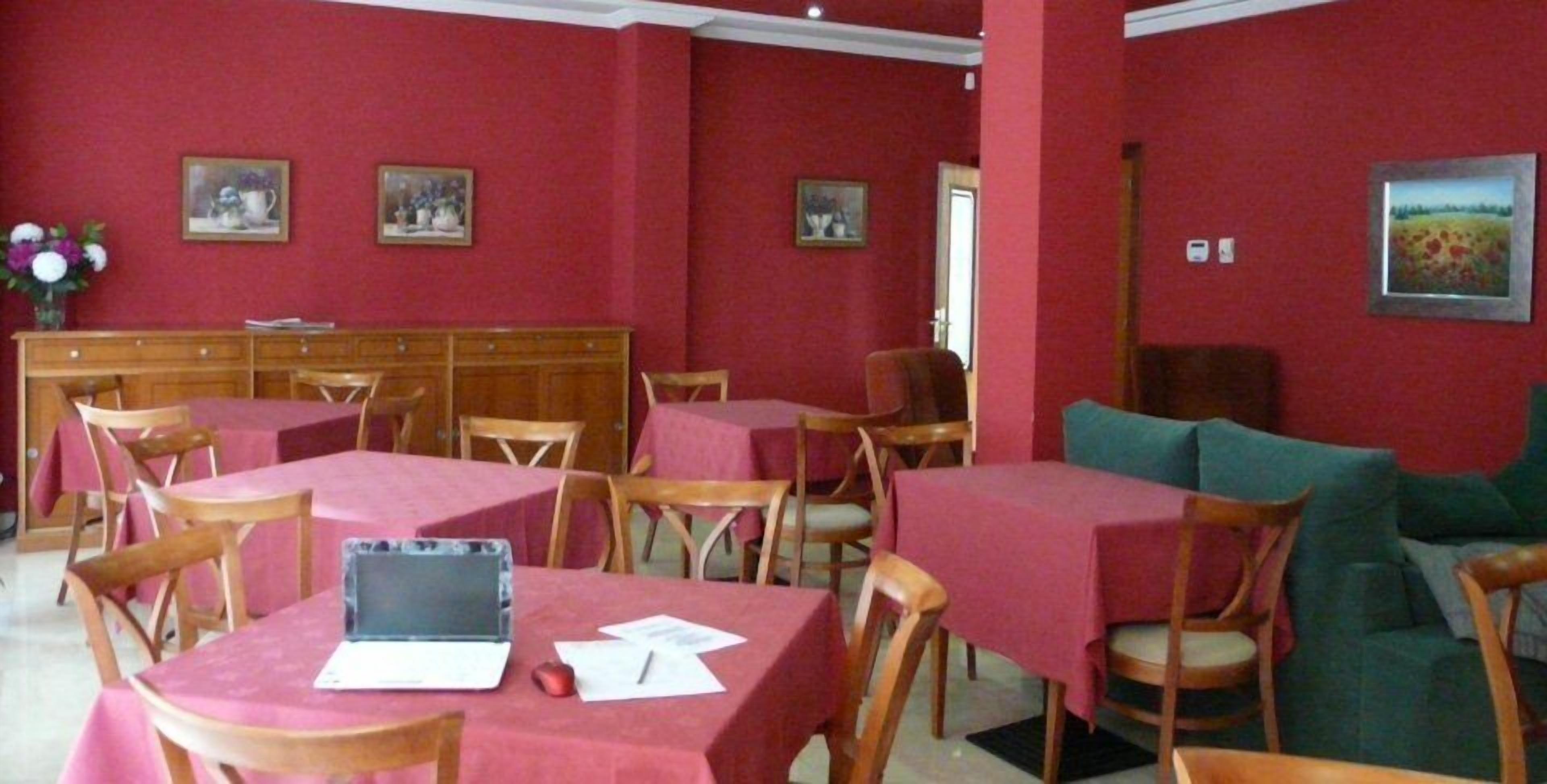 A dining area with red walls, wooden tables, and chairs. There are framed pictures on the walls and a laptop on one table.