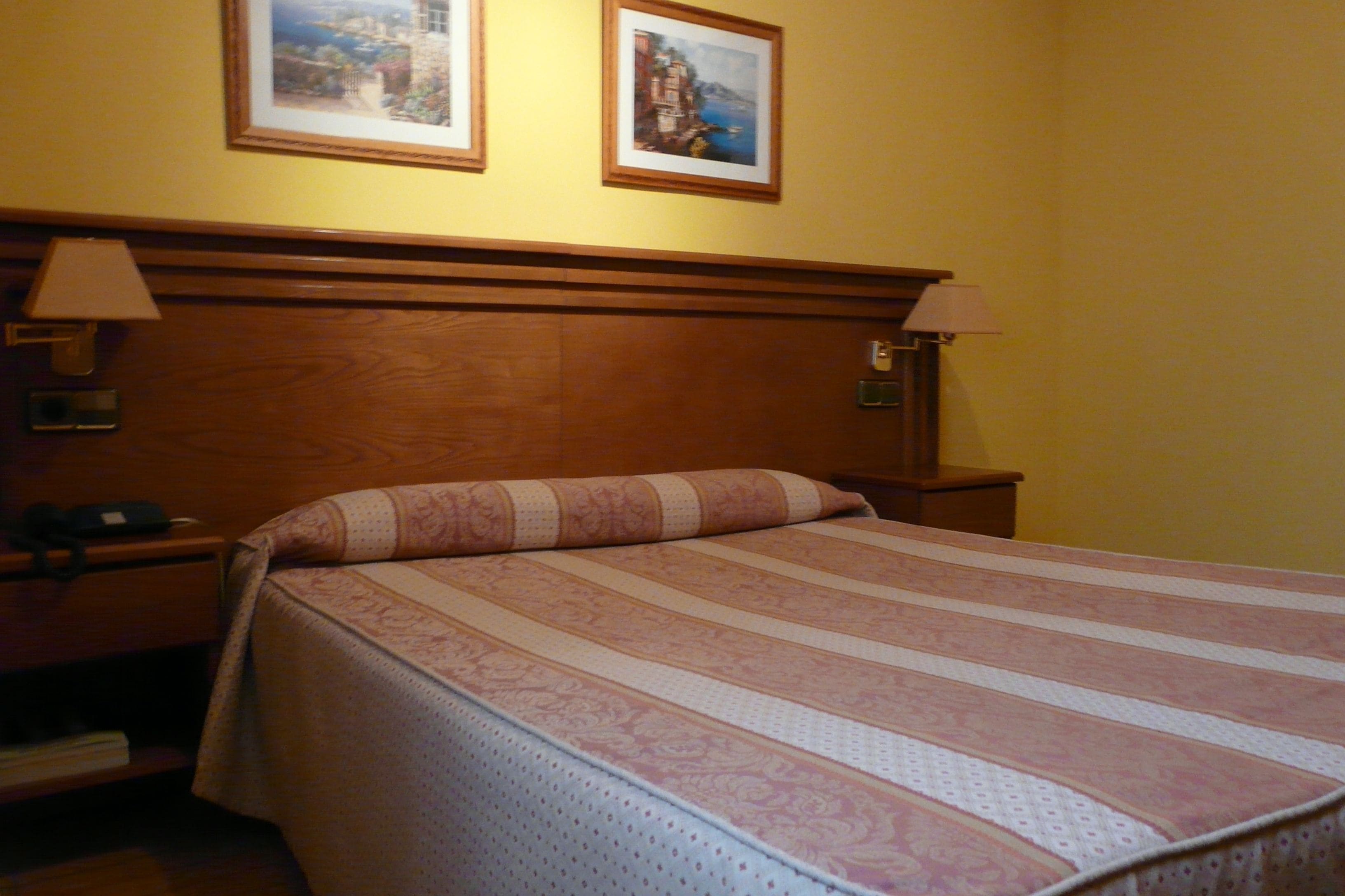 A hotel room with a wooden headboard, two bedside lamps, a bed with a striped bedspread, and two framed pictures on the wall.