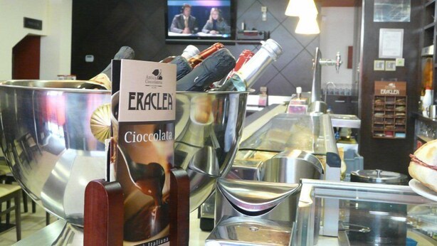 Una zona de bar con un gran contenedor metálico que contiene una botella de Eraclea Cioccolata. Hay varias botellas y vasos sobre el mostrador, y un televisor está montado en la pared al fondo.