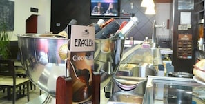 A bar area with a large metal container holding a bottle of Eraclea Cioccolata. There are various bottles and glasses on the counter, and a television is mounted on the wall in the background. - Hotel IDH Angel (Oviedo)