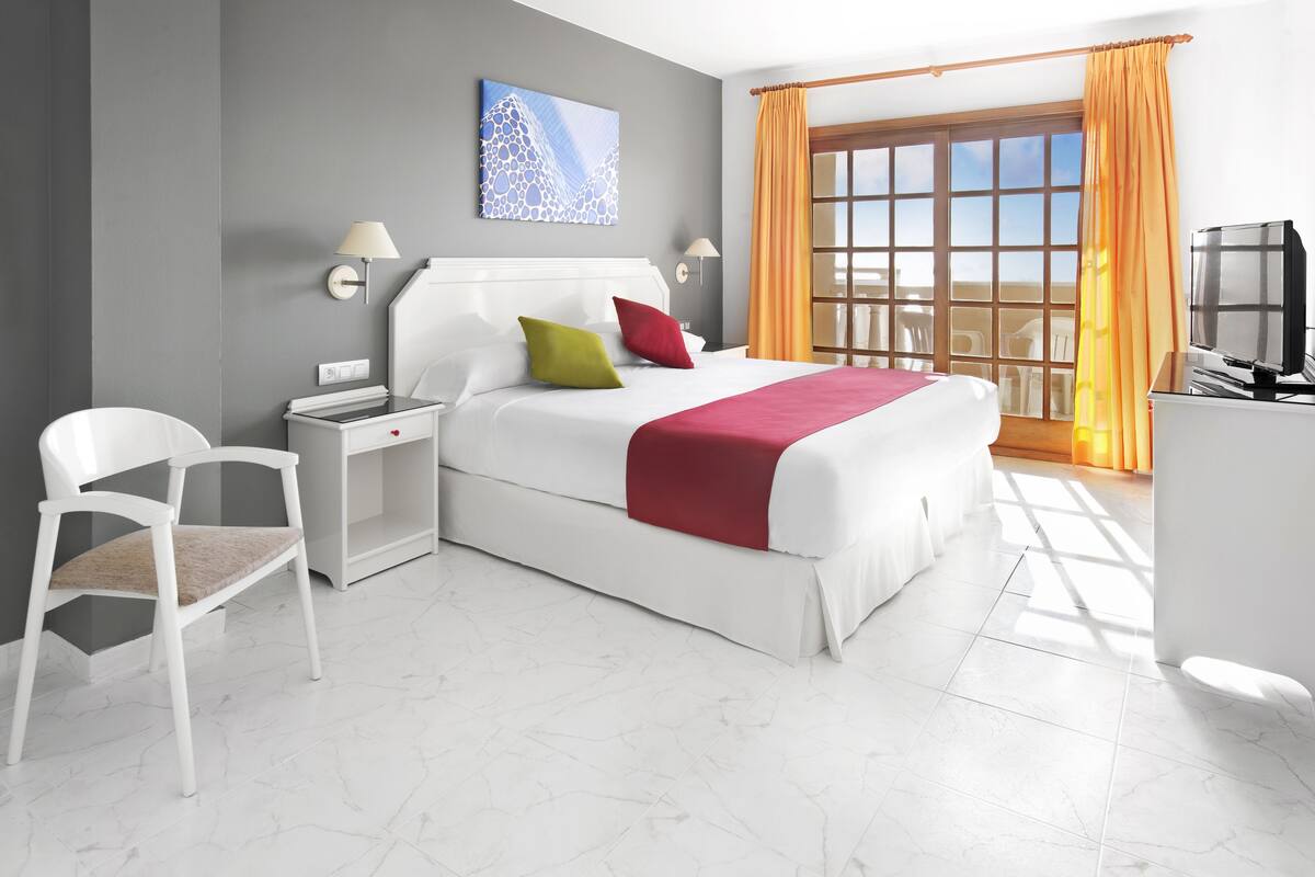 Habitación de hotel moderna con una cama grande, una silla blanca, un escritorio con televisor y un balcón con cortinas naranjas.