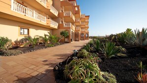 Un hotel de varios pisos con balcones, un paseo cubierto de baldosas y jardines ajardinados con diversas plantas.