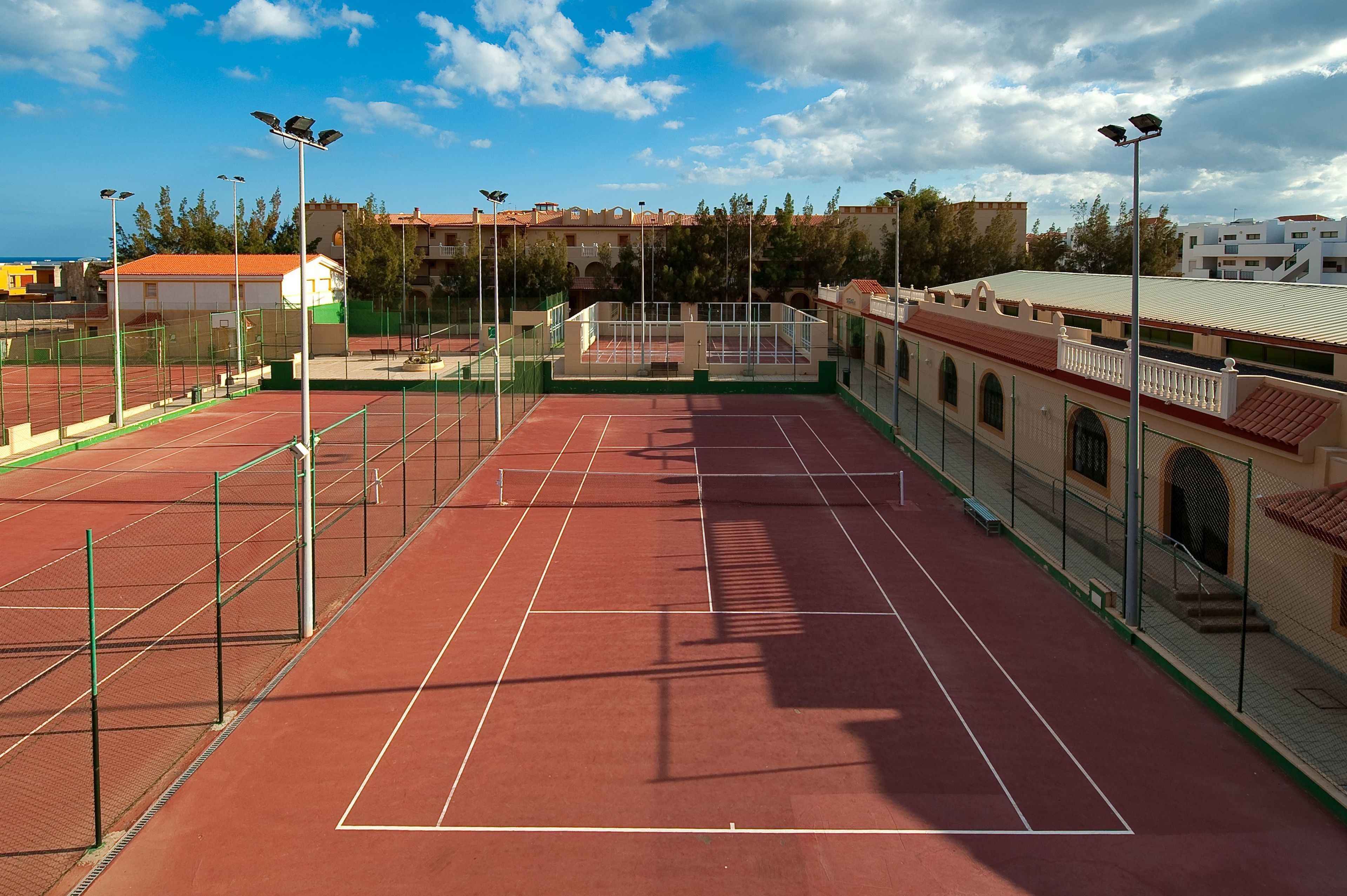 A tennis court with a red surface, surrounded by a green fence, and adjacent buildings with red roofs.