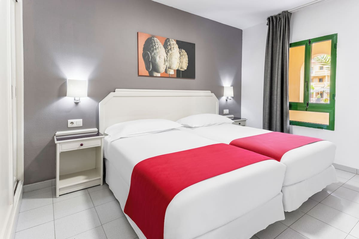 A double bed with a red blanket, a nightstand with a lamp, a wall-mounted artwork, and a window with a green frame.