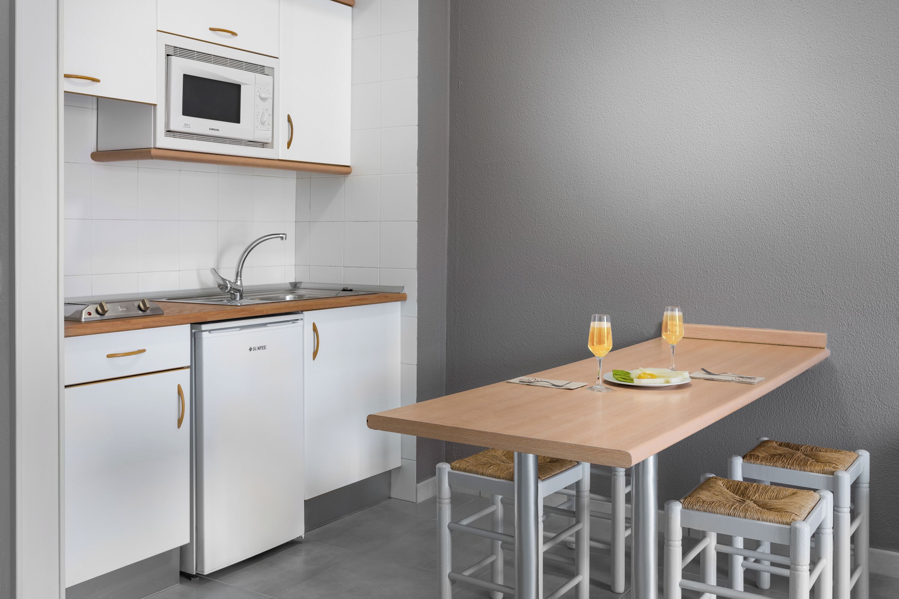 A compact kitchen with a microwave, sink, and white cabinetry. There is a wooden countertop and a table with two chairs.