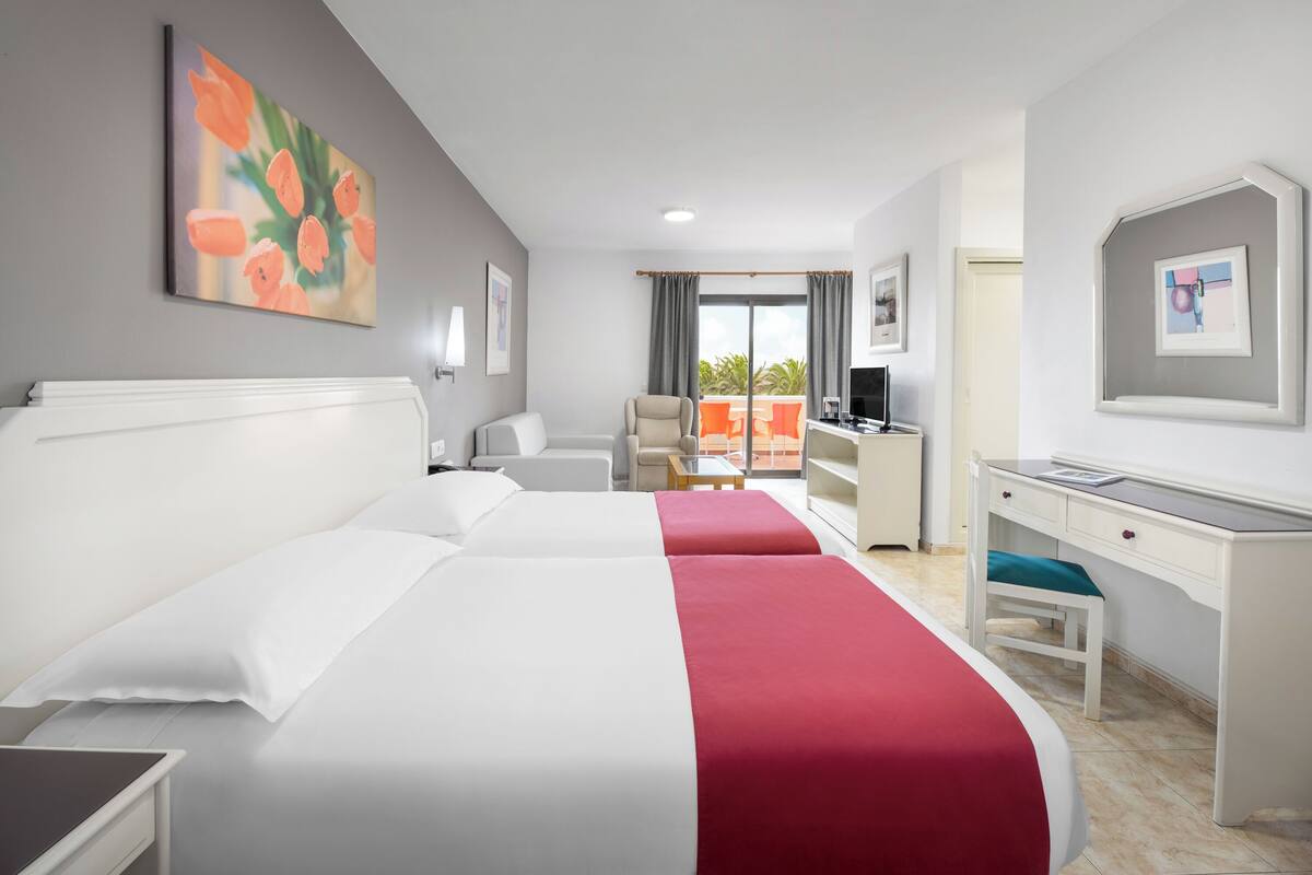 A hotel room with two beds, a desk, a chair, a balcony with a view, and artwork on the walls.
