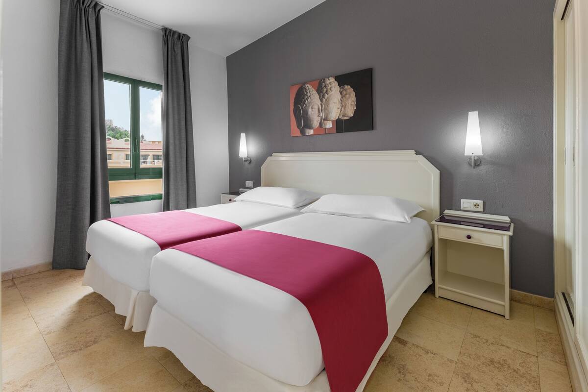A double bed with a white and pink bedspread, two bedside tables with lamps, a painting on the wall, and a window with curtains.
