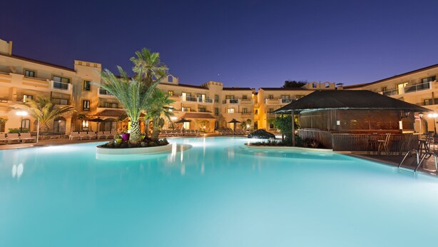 Ein Swimmingpool mit Liegestühlen und Sonnenschirmen, umgeben von Hotelgebäuden mit Balkonen und Sitzbereichen im Freien.