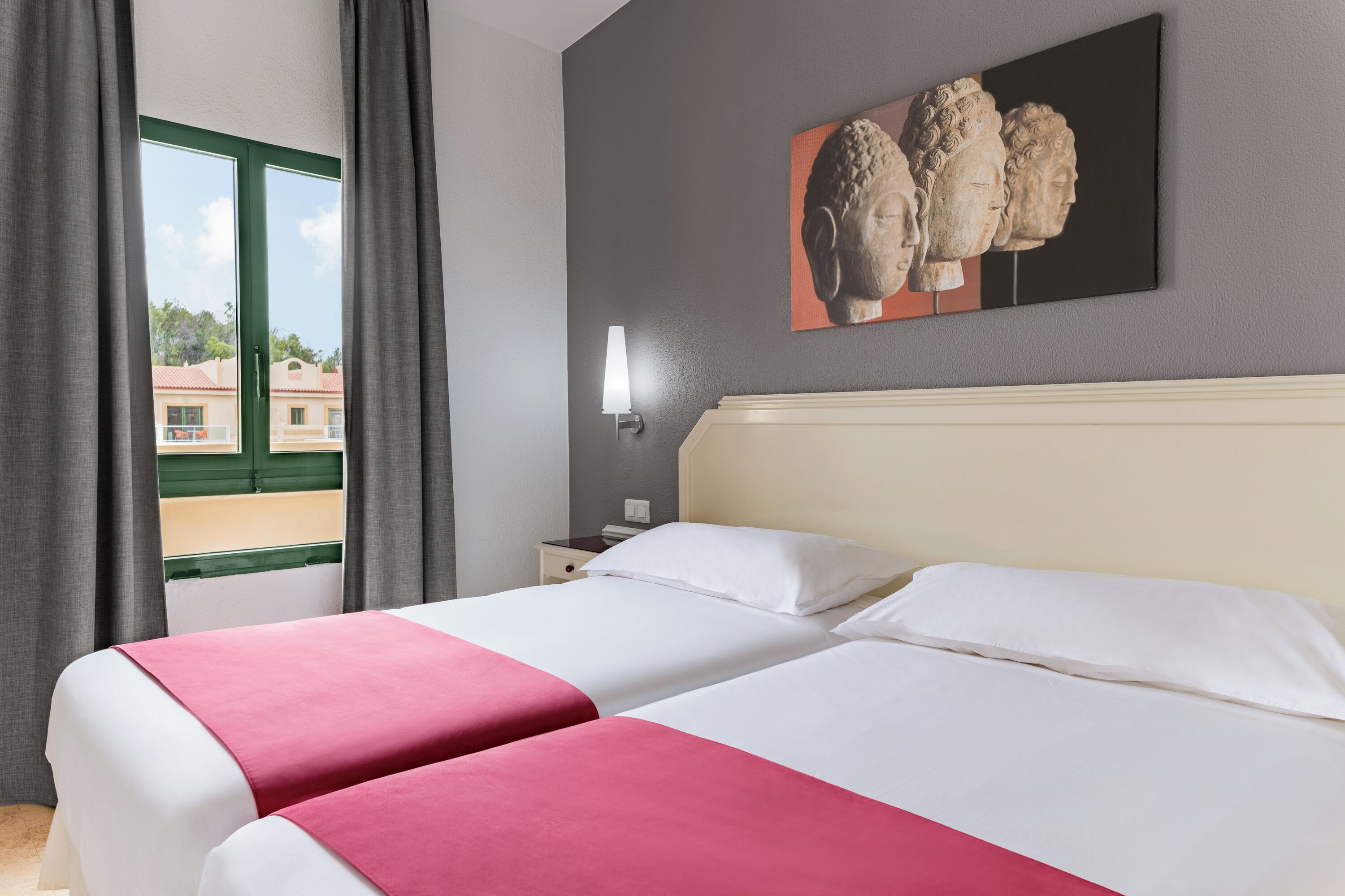 A hotel room with two beds, a window with curtains, a nightstand, a wall-mounted lamp, and a painting of busts.