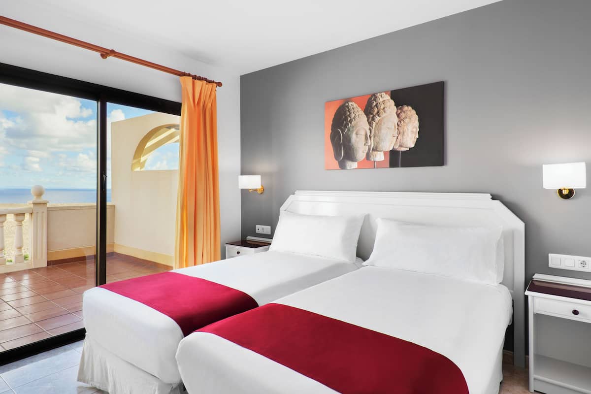 A hotel room with two beds, a balcony, a painting, and a view of the sea.