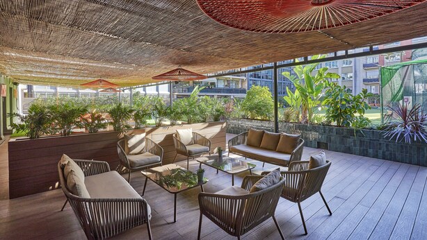 A modern outdoor lounge area with wicker furniture, a glass table, and potted plants. - The Moods Oasis (Barcelona)