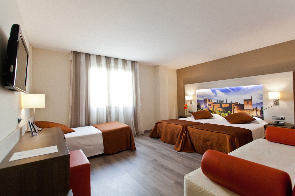 Habitación de hotel con dos camas, un escritorio, un televisor y un amplio ventanal con cortinas.