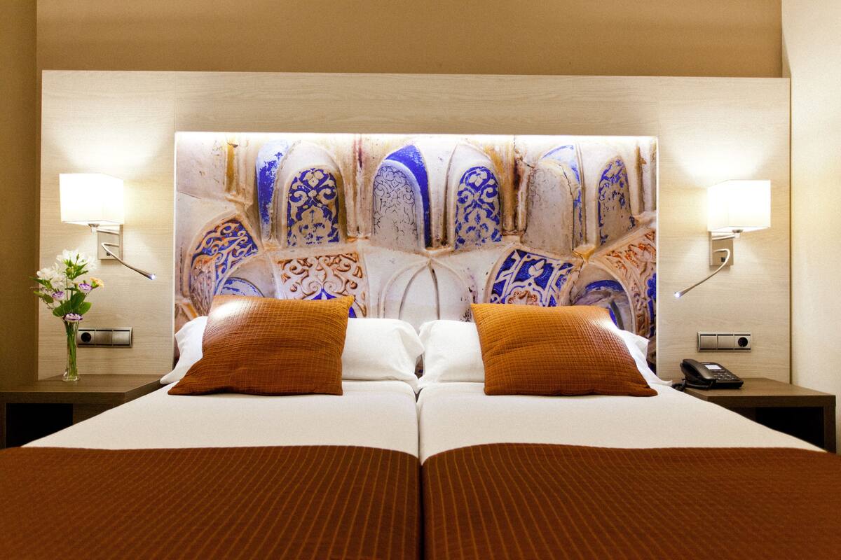 Una cama doble con un cabecero decorativo, lámparas de noche y un jarrón con flores.