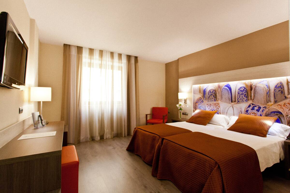 Habitación de hotel con cama, escritorio, silla, televisor y ventana con cortinas.